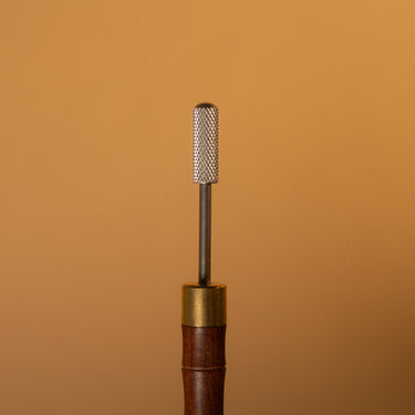 A paint roller tool with a textured metal tip and sturdy wooden handle, used for applying paint to leather surfaces, laid out on a solid light brown background.